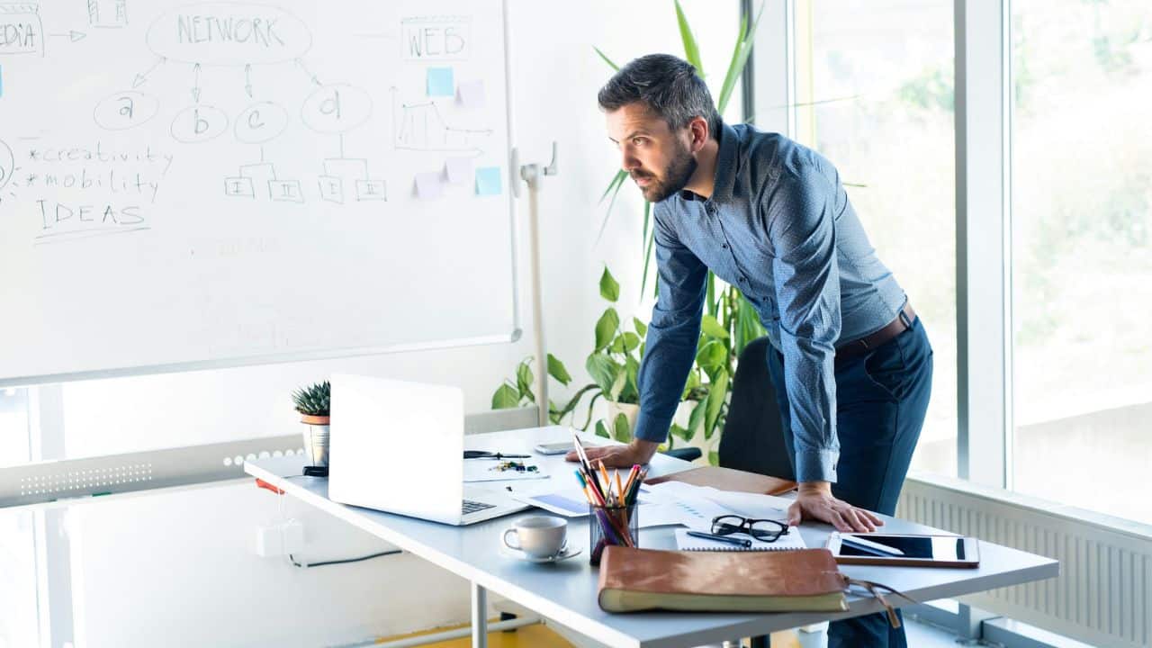 A professional man leans on a desk in an office, looking at a whiteboard with various diagrams.