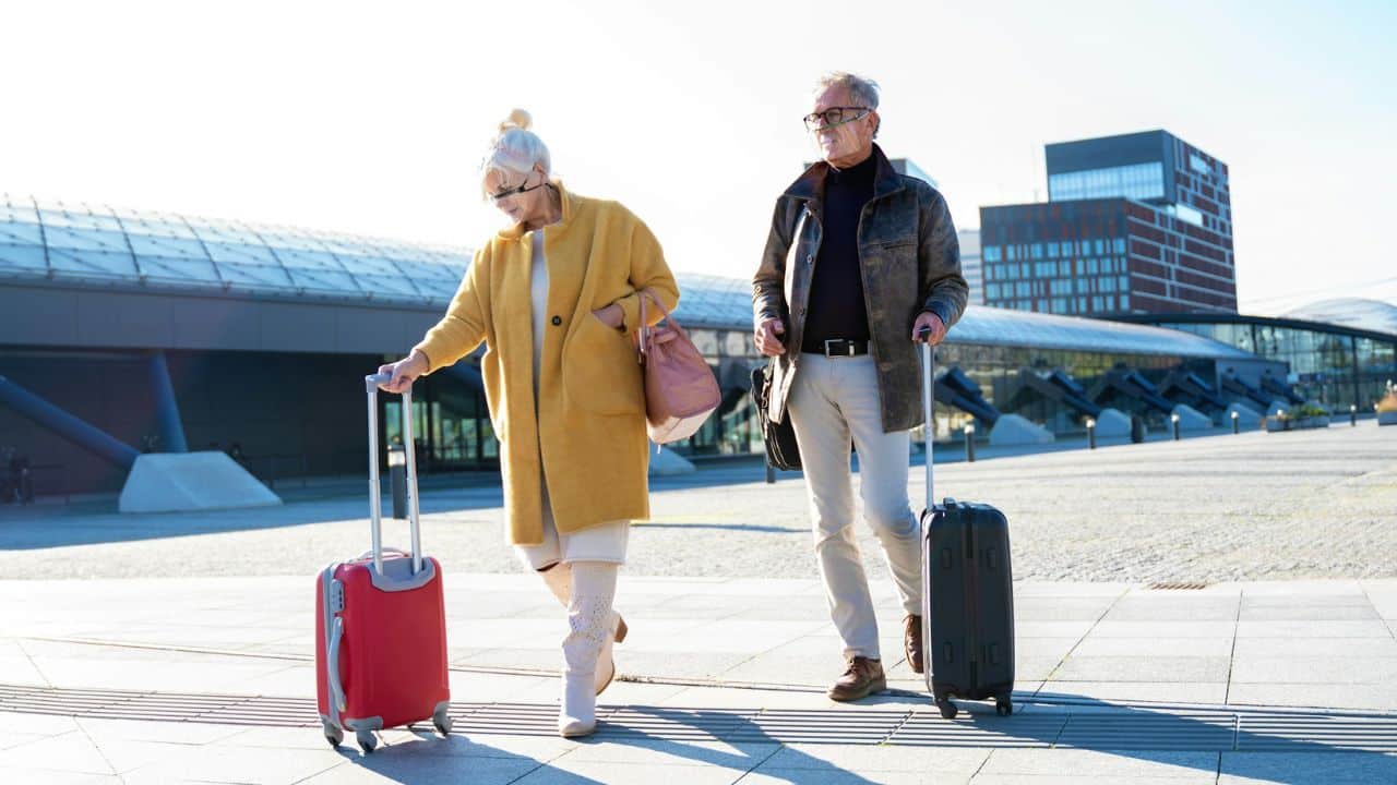 A fashionable elderly couple pulls their suitcases while walking in front of a modern building.