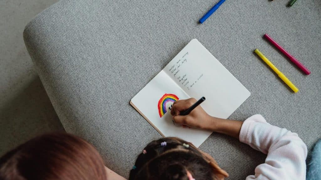 A child draws a rainbow in a notebook with colored markers.