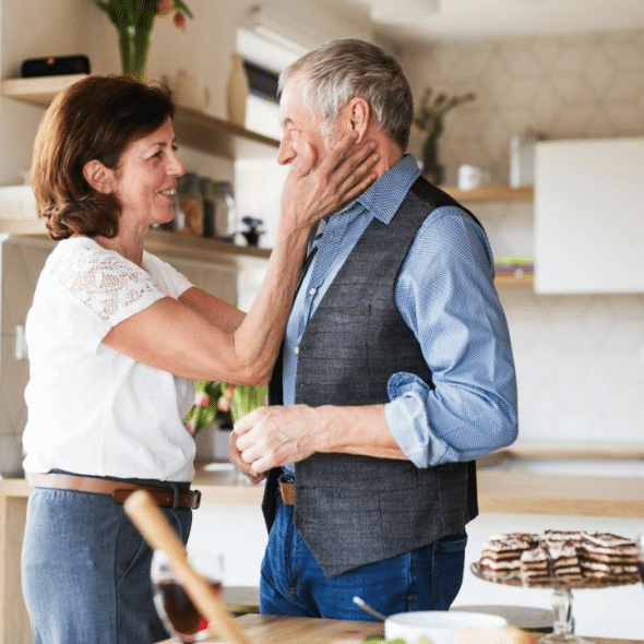 A happy, older woman wearing a white shirt lovingly touches the face of a smiling, older man.