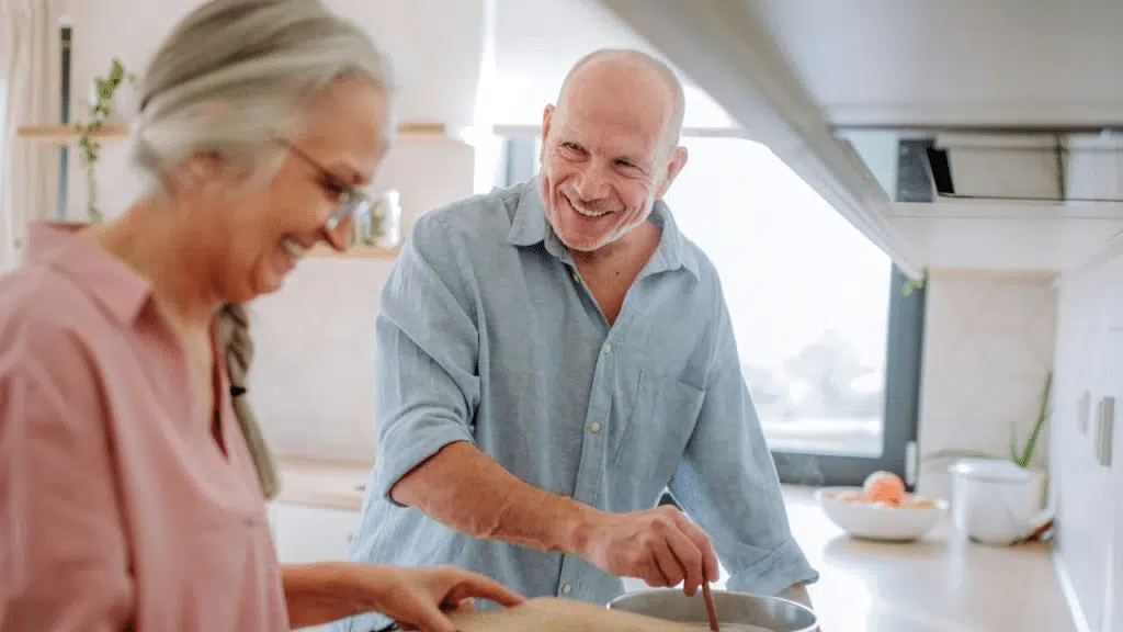 A joyful elderly couple smiles while cooking together in a bright kitchen.