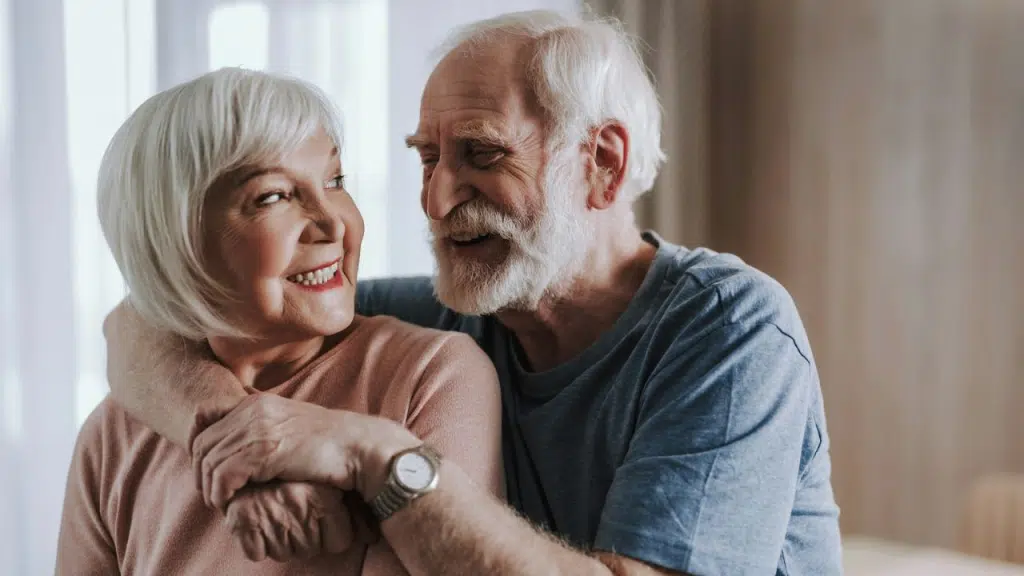 A cheerful elderly couple with white hair embraces, looking affectionately at each other.
