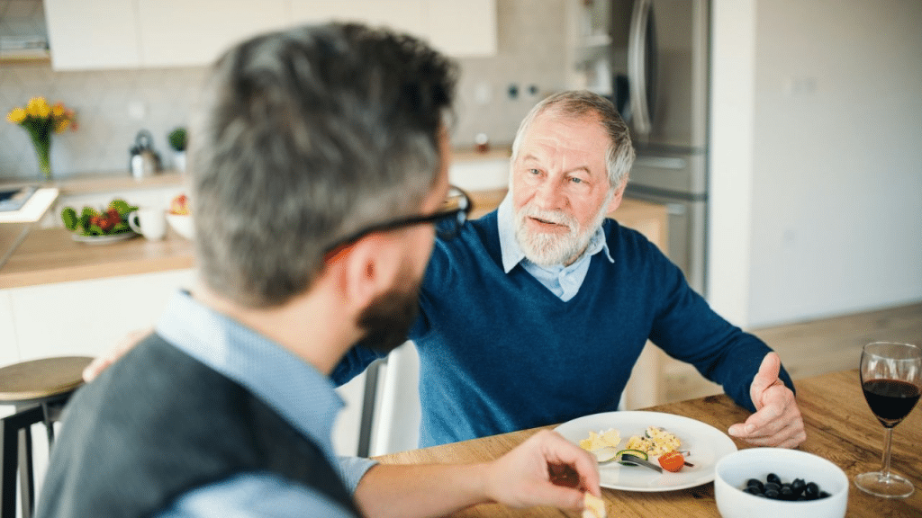 An older man talks and gestures while sitting at a kitchen table.