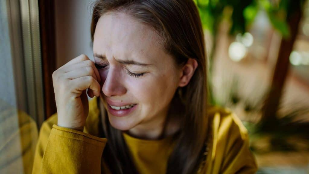 A woman in a yellow sweater cries by a window.