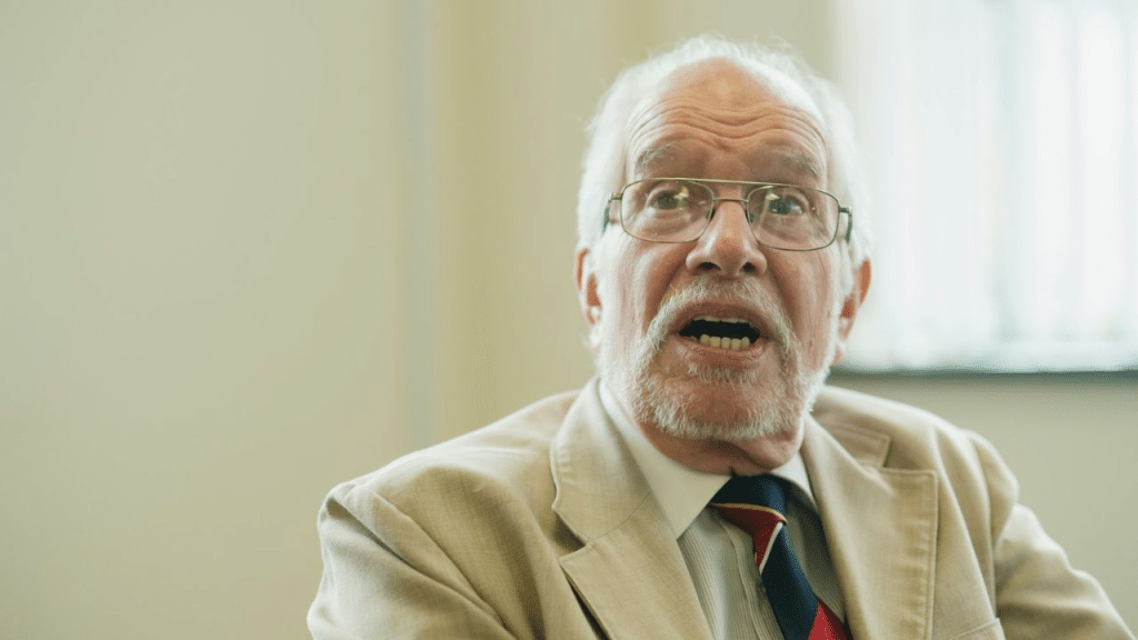 A surprised elderly man with glasses and a white beard talks, wearing a beige jacket and a tie.