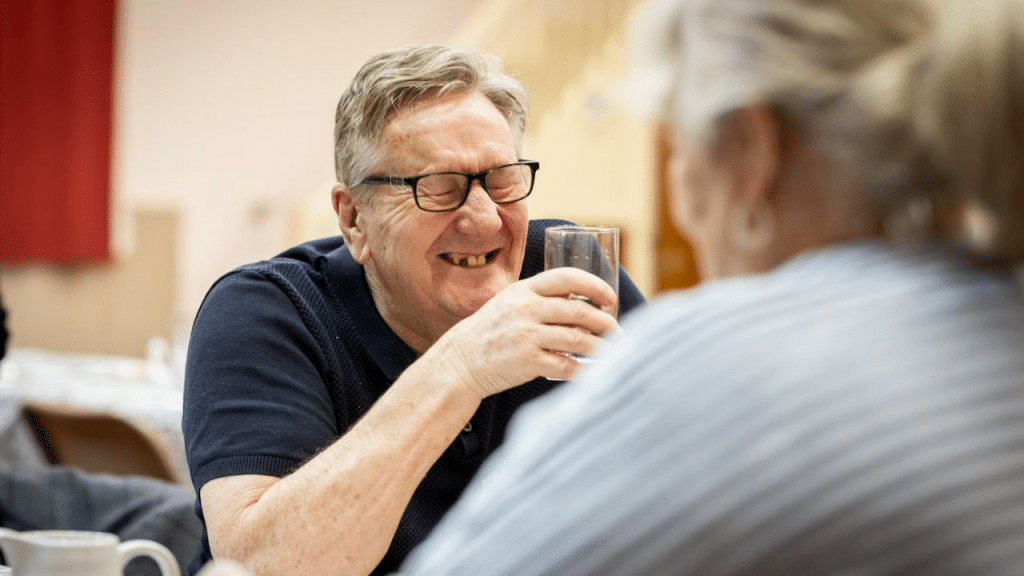 A man with glasses and a missing tooth laughs while holding a glass of water.