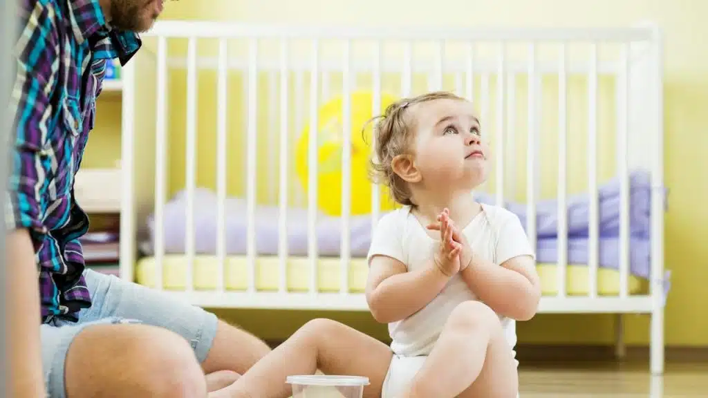 A toddler sits on the floor near a crib looking upward.