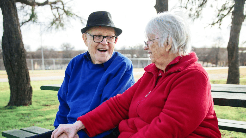 A joyful elderly couple sits on a park bench, holding hands and laughing.