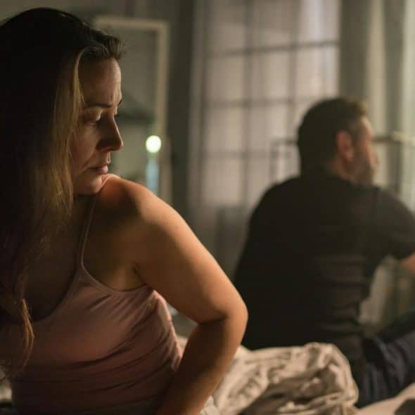 A woman looking upset while sitting on a bed with a man facing away near a window.