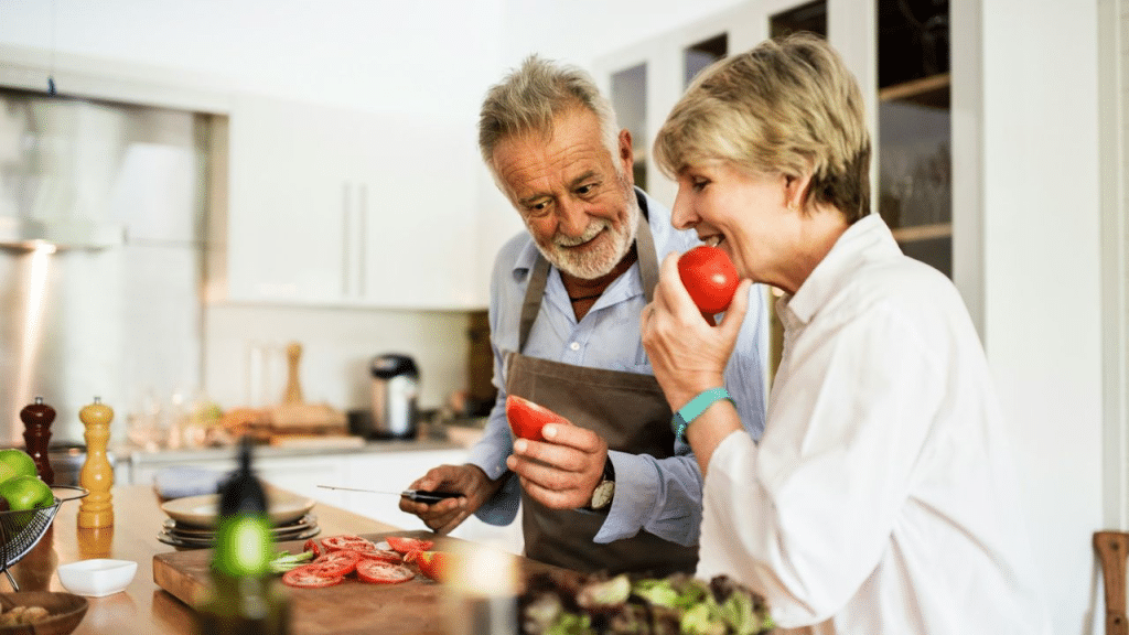 A cheerful, gray-haired couple is happily cooking and preparing a meal together in a bright kitchen.