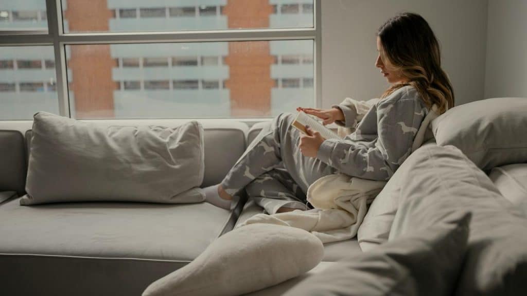 A woman in pajamas reading a book on a cozy couch near a window.