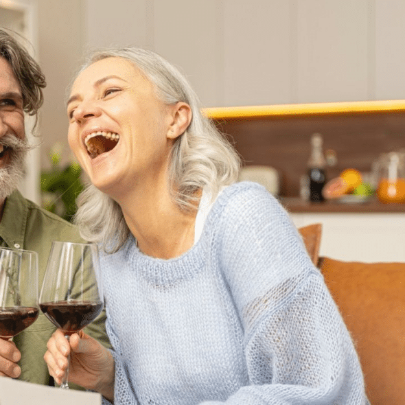 A laughing elderly couple with gray hair is toasting with glasses of red wine.