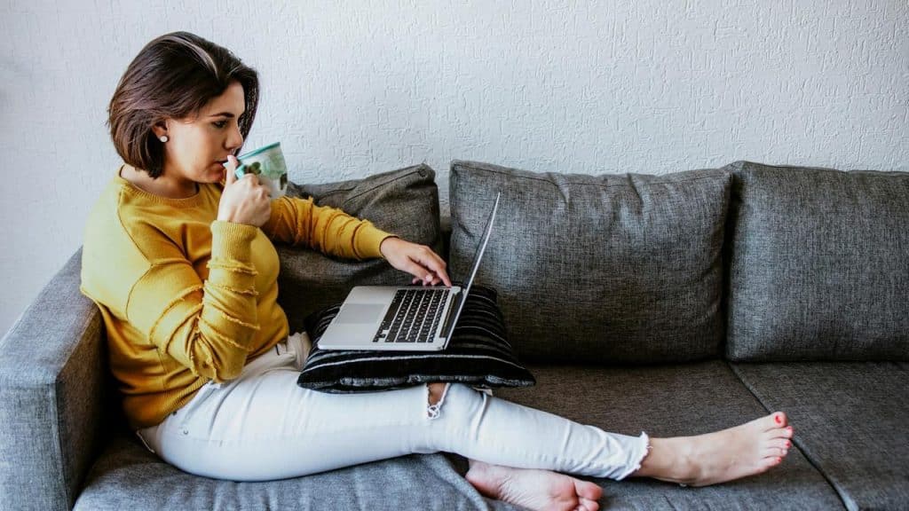 A woman sitting on a couch drinking coffee while using a laptop.