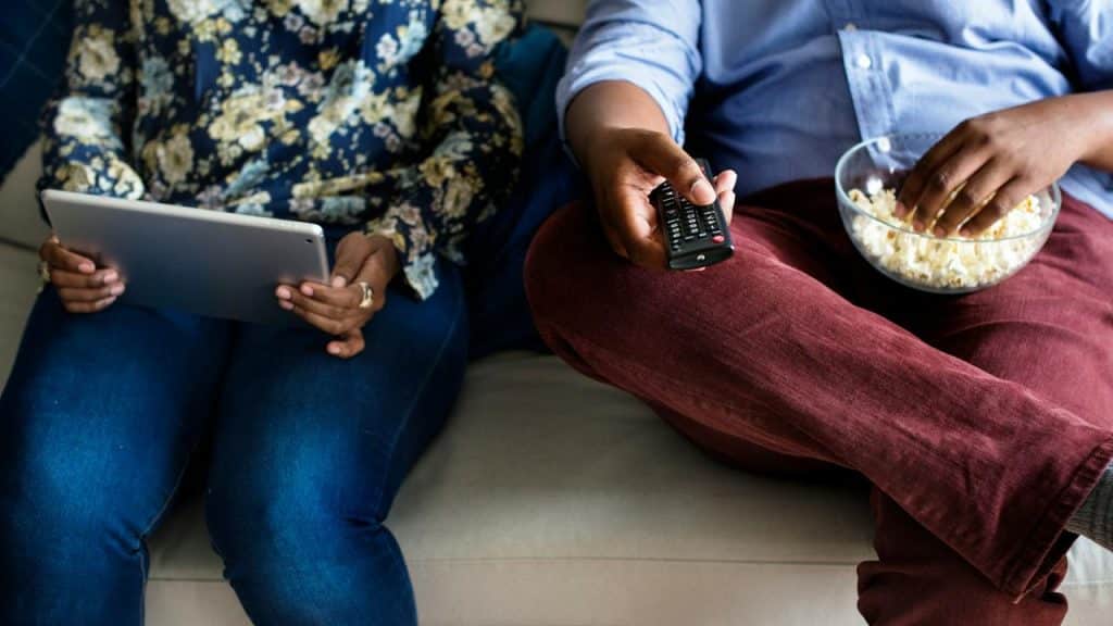 A couple on a couch with a tablet, popcorn, and a remote.