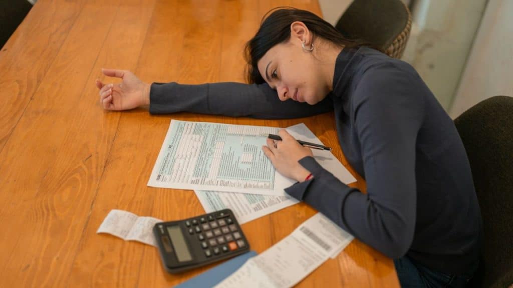 A frustrated woman with her head on a table works on paperwork.