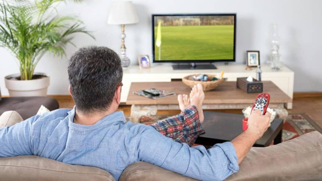 A man in a blue shirt and plaid pants relaxes on a couch, holding a remote control.