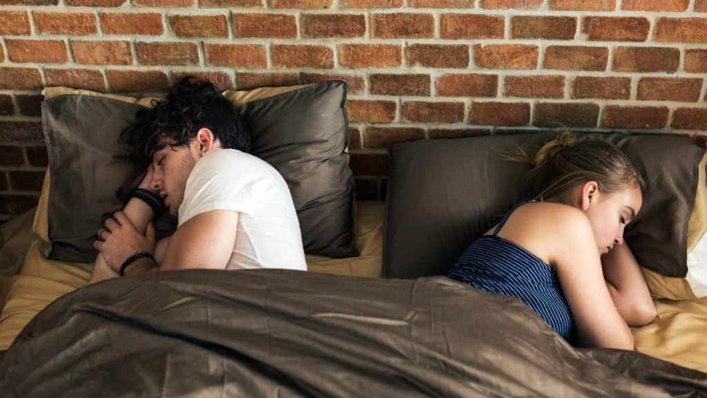 A man and woman lie in bed back-to-back, facing away from each other.