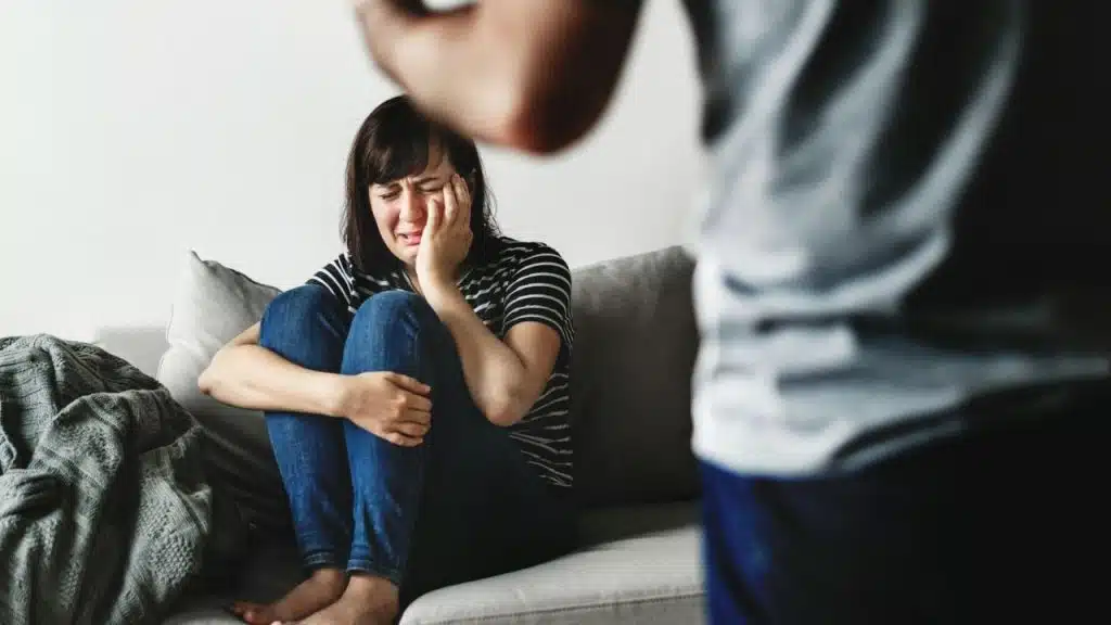 A distraught woman sits crying on a couch with a person out of focus in the foreground.