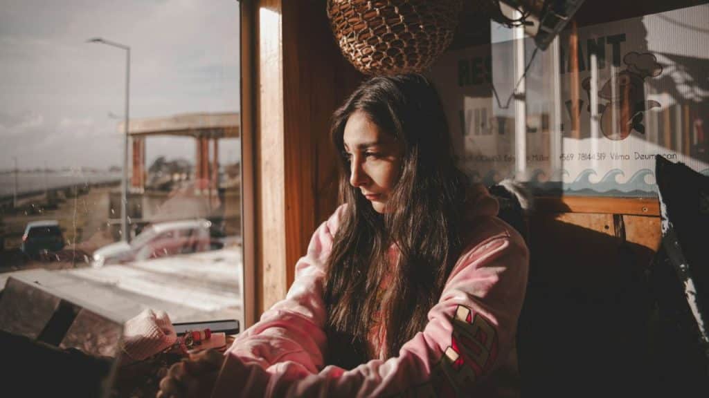 A sad young woman with long dark hair looks out a window in a cafe.