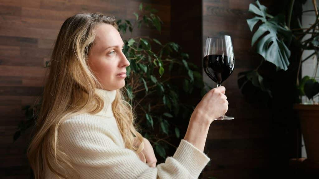 A woman in a cream-colored sweater holds a glass of red wine, looking into the distance.