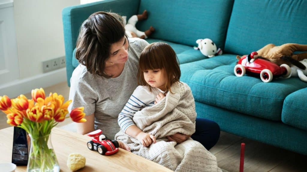 A smiling mother comforts a sad child wrapped in a blanket on the floor.