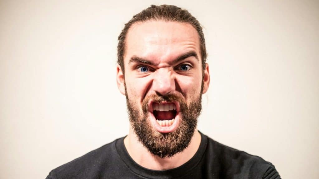 A bearded man with wide blue eyes grimaces and bares his teeth.