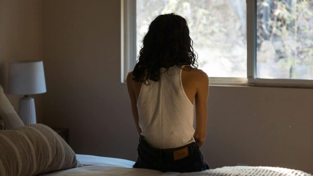 A woman with her back to the camera sits on a bed and looks out a window.