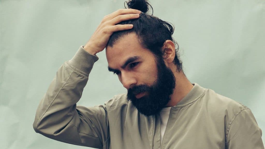 A man with a beard and hair bun holds his head, looking down.