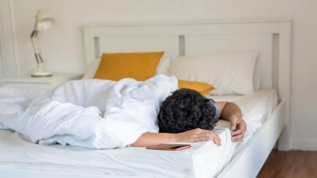 A person lies facedown in bed, with a smartphone on the mattress next to them.