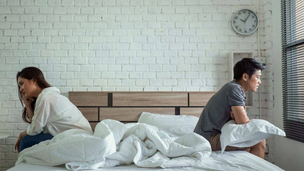 A man and woman sit separately on a bed with their backs to each other, looking upset.