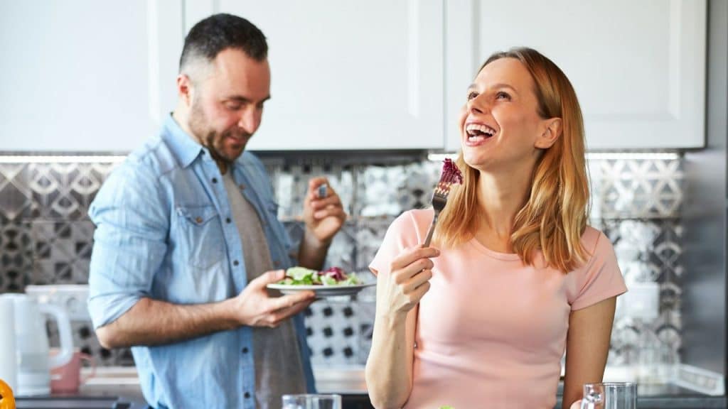 A happy woman in a pink shirt laughs while eating a salad, and a man in a denim shirt stands behind her.