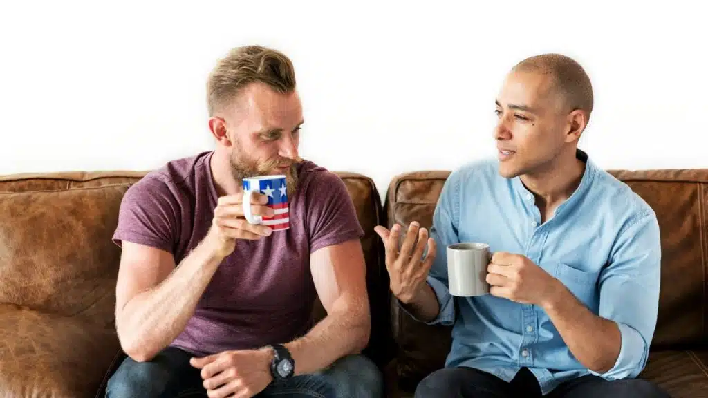 Two men sit on a brown leather couch, drinking from mugs and talking to each other.