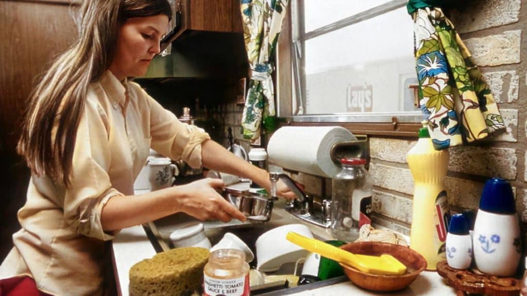 A woman with long brown hair washes dishes in a cluttered kitchen.