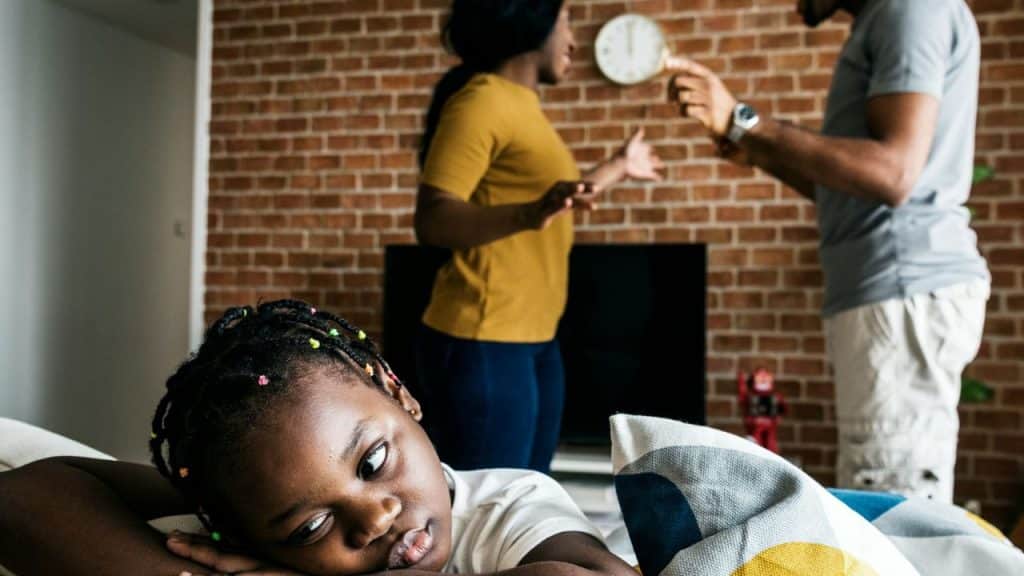 A little girl with braids looks sad as her parents argue in the background.