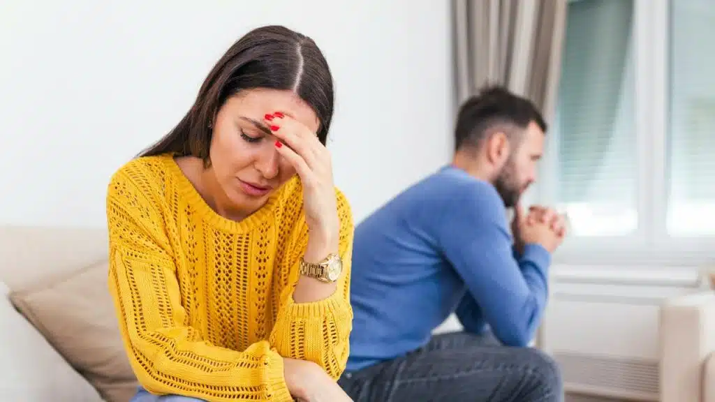 A worried woman with her hand on her forehead sits in the foreground, with a man out of focus in the background.