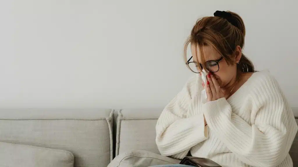 A woman in a white sweater sitting on a couch blowing her nose.