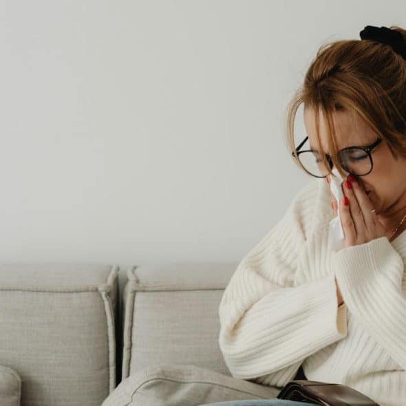 A woman in a white sweater sitting on a couch blowing her nose.