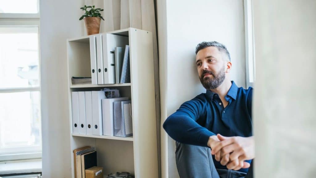 A sad man with a beard sits on the floor, looking into the distance.