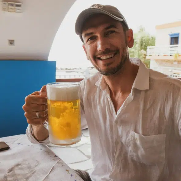 A joyful man in a white shirt and baseball cap holds a large mug of beer.