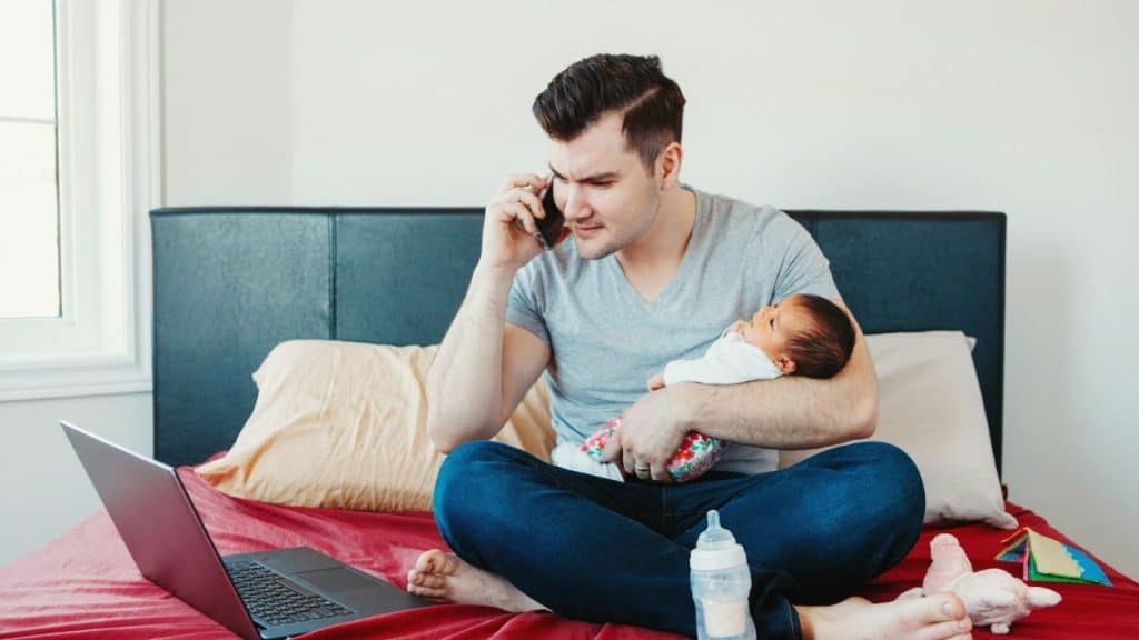 A man holds a baby while talking on the phone and looking at a laptop.