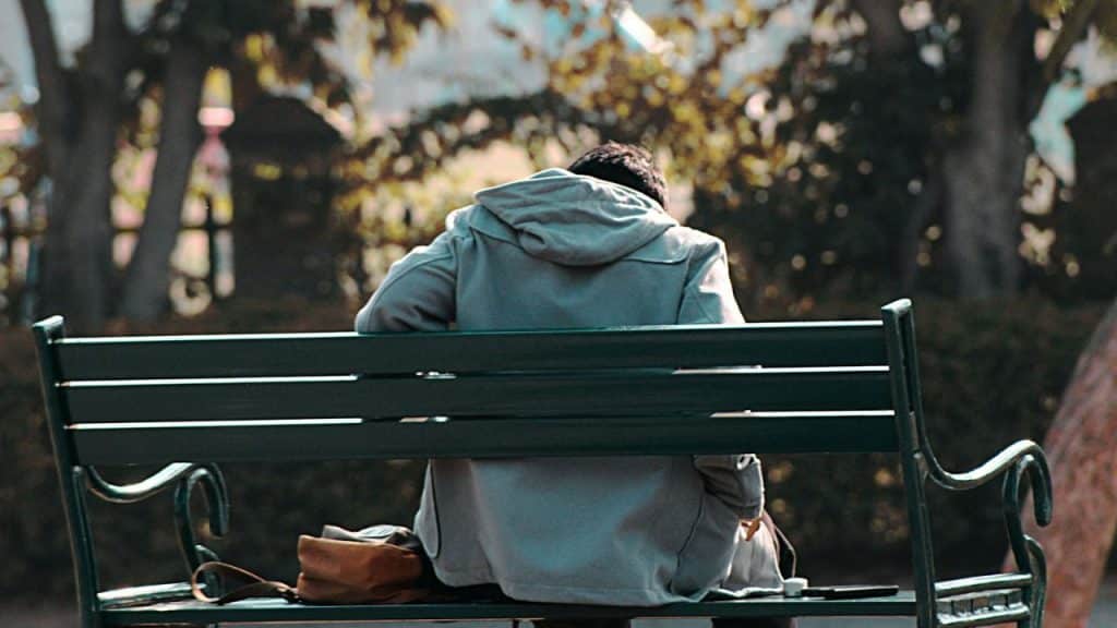 A person in a gray hoodie sits alone on a park bench, facing away from the camera.