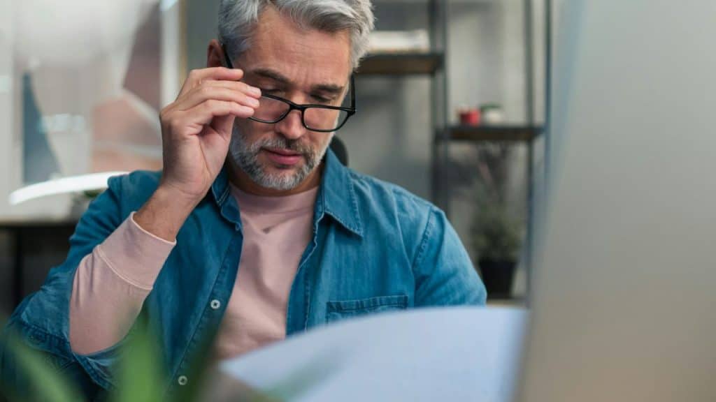 A focused man with gray hair and a beard looks at documents, adjusting his glasses.