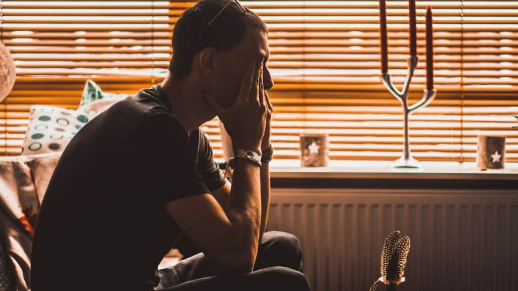 A sad man in a dark t-shirt sits with his hands covering his face, looking down.