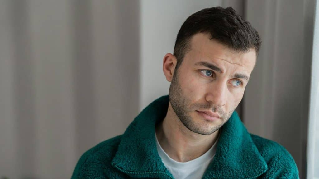 A sad man in a dark t-shirt sits with his hands covering his face, looking down.