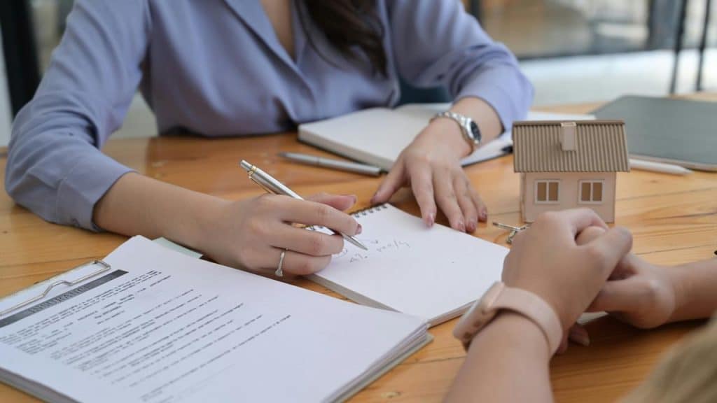 A person signing paperwork at a desk with a small house model.