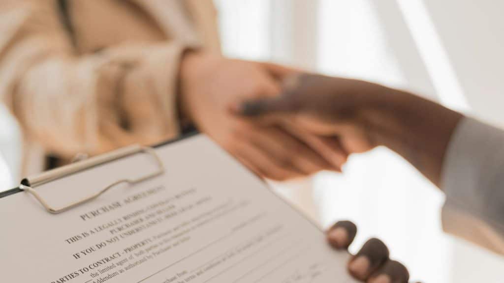 A contract agreement on a clipboard with two people shaking hands in the background.