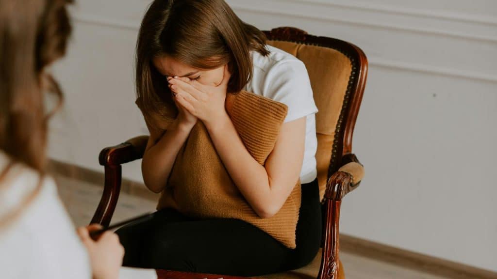 A woman sitting in a chair covering her face with her hands while holding a pillow.