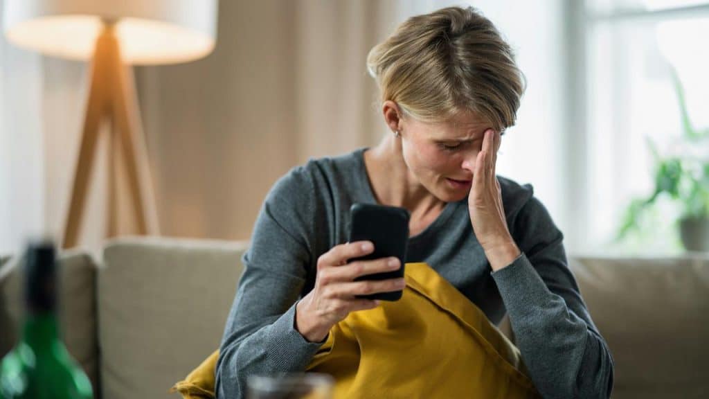 A woman sits on a couch holding a phone and looking distressed.