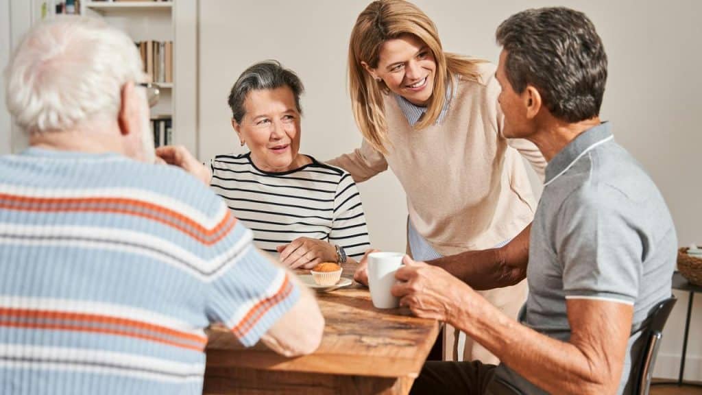 A group of older adults sit at a table talking with a smiling woman.