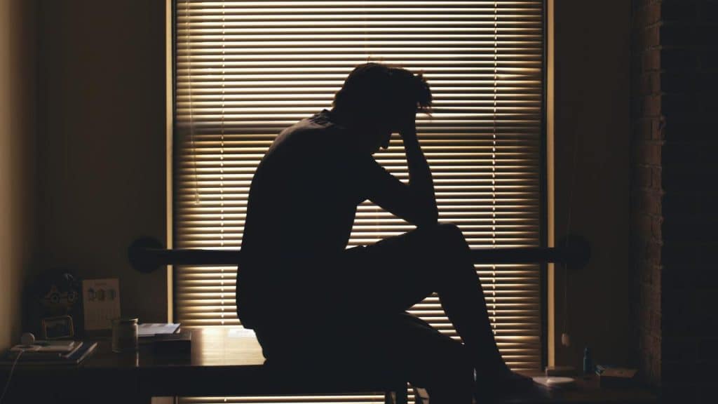 A man sits in silhouette with his head in his hand by a window.